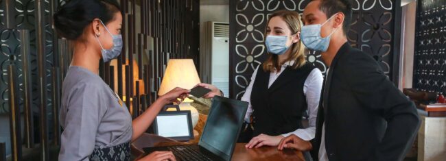 Couple and receptionist at counter in hotel wearing medical masks.